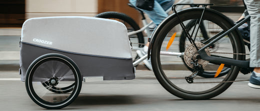 Man rides bike one a road with Croozer Cargo trailer in stone grey; daughter cycles beside him.